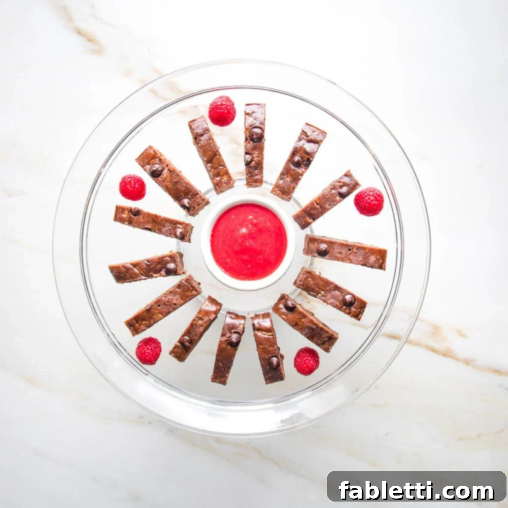 Secret Garden Chocolate Brownies 6 Thinly sliced chocolate zucchini brownies arranged in a circle around a small bowl of vibrant raspberry sauce, inviting a dip.