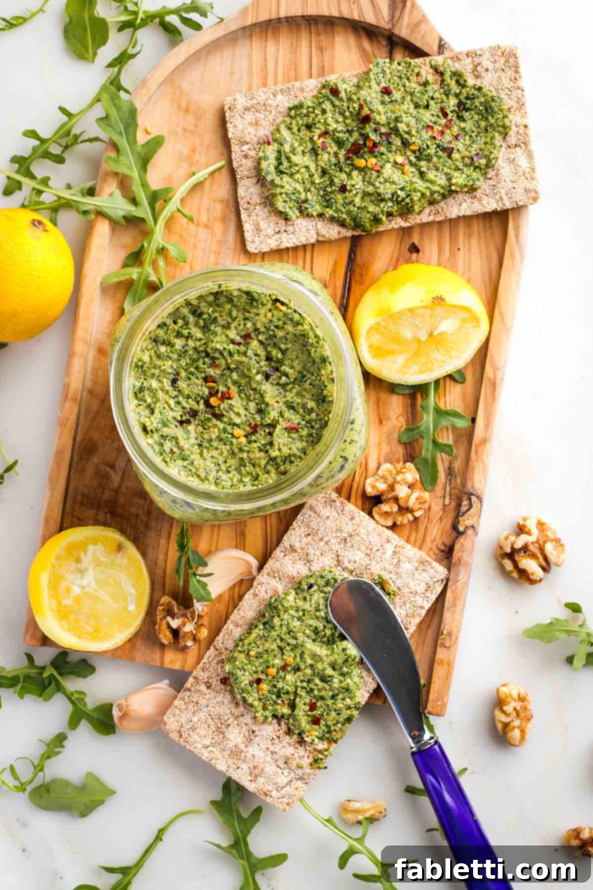 Serving Walnut Pesto with Crackers A charming scene featuring a glass jar of freshly made walnut arugula pesto. It is thoughtfully surrounded by whole lemons, walnuts, fresh arugula leaves, garlic cloves, and several long, crunchy crackers generously spread with the vibrant green pesto, all ready for a delicious tasting.