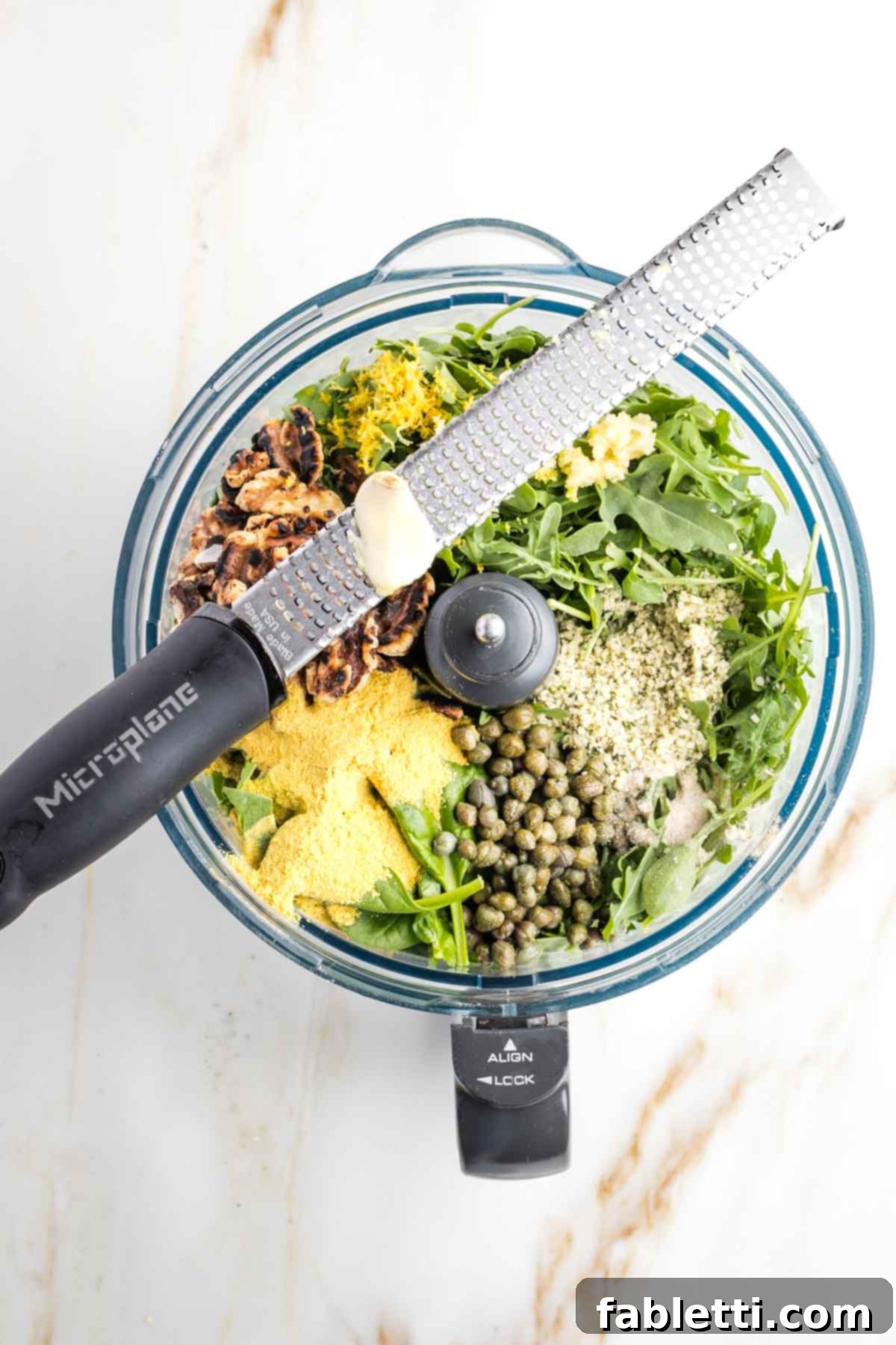 Pesto Making Pro Tip: Zesting Lemon Directly Into Processor A close-up photograph of a hand expertly zesting a fresh lemon directly over the open bowl of a food processor, illustrating an efficient culinary technique that saves time and minimizes clean-up during pesto preparation.