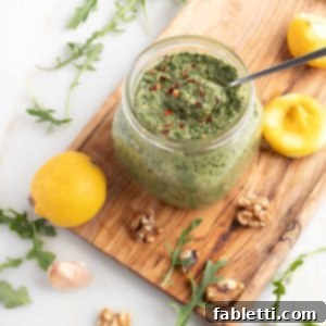 Vegan Walnut Pesto with Arugula Recipe A glass jar of freshly made vegan walnut pesto with arugula, beautifully garnished with a sprinkle of crushed red pepper for a touch of heat. The jar is surrounded by fresh lemons, walnuts, vibrant arugula leaves, and aromatic garlic cloves, all ready to be incorporated or enjoyed with the pesto.