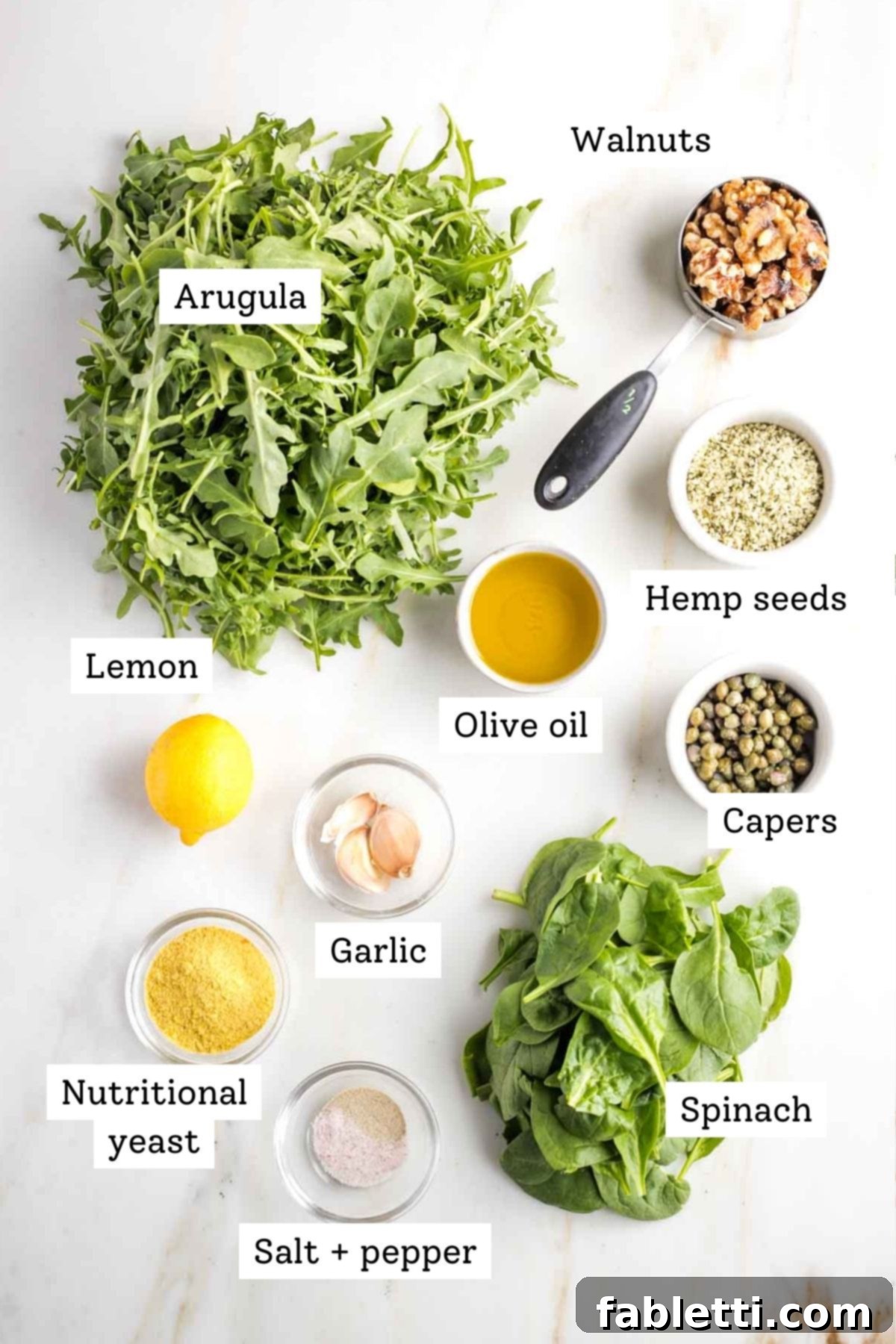 Walnut Pesto Ingredients List A beautifully arranged flat lay of all labeled ingredients for making walnut pesto: fresh arugula, crunchy walnuts, protein-rich hemp seeds, golden extra virgin olive oil, briny capers, a bright lemon, pungent garlic, fresh spinach, savory nutritional yeast, and essential salt and white pepper.