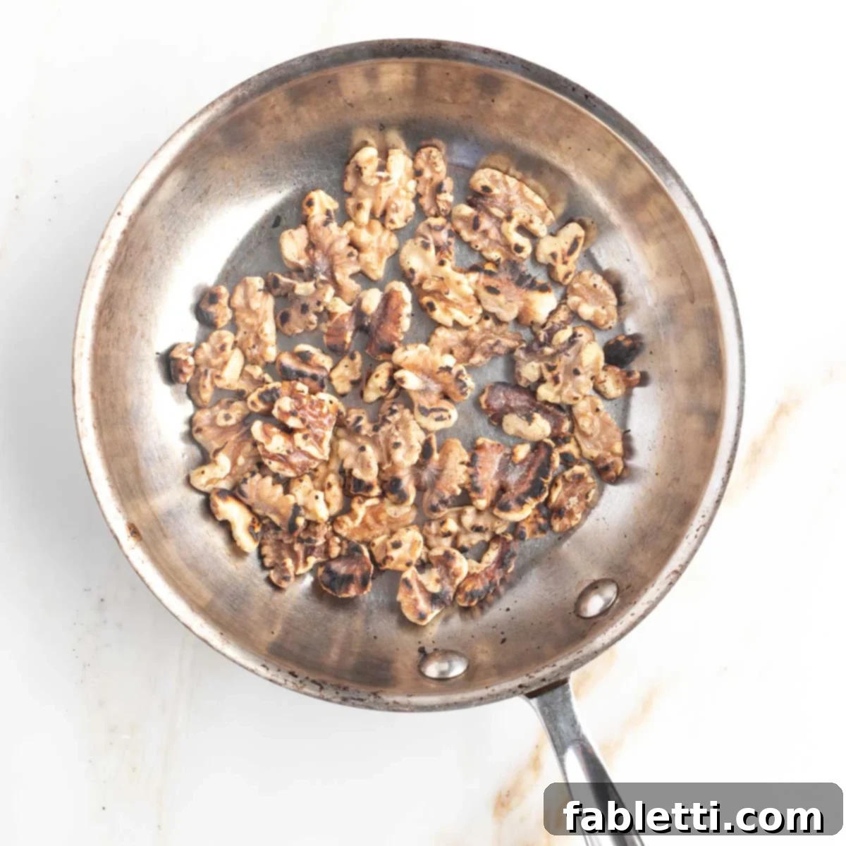 Toasting Walnuts for Pesto Raw walnuts toasting gently in a dry stainless steel pan over medium heat. The nuts are beginning to turn golden and release their aromatic oils, a crucial step for enhancing their flavor for the pesto.