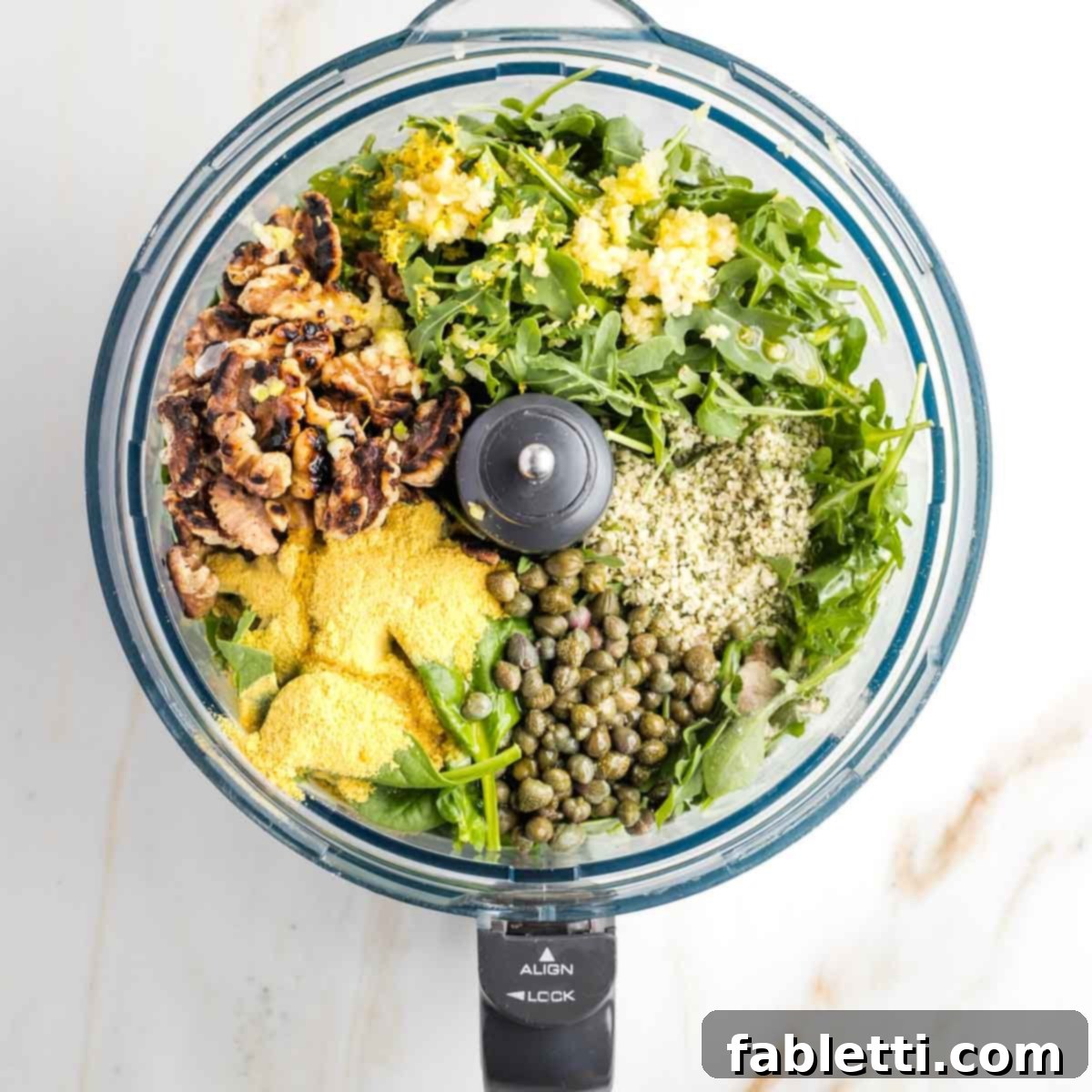 Pesto Ingredients in Food Processor The spacious bowl of a food processor filled with a colorful array of fresh greens including arugula and spinach, along with toasted walnuts, briny capers, protein-rich hemp seeds, aromatic lemon zest, and garlic cloves, all poised for blending into a delicious pesto.