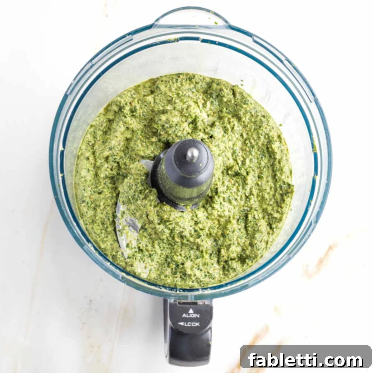 Blended Walnut Arugula Pesto The food processor bowl now holds a perfectly blended, creamy, and vibrant bright green pesto. Its smooth texture indicates thorough processing to achieve the desired consistency, ready for serving.
