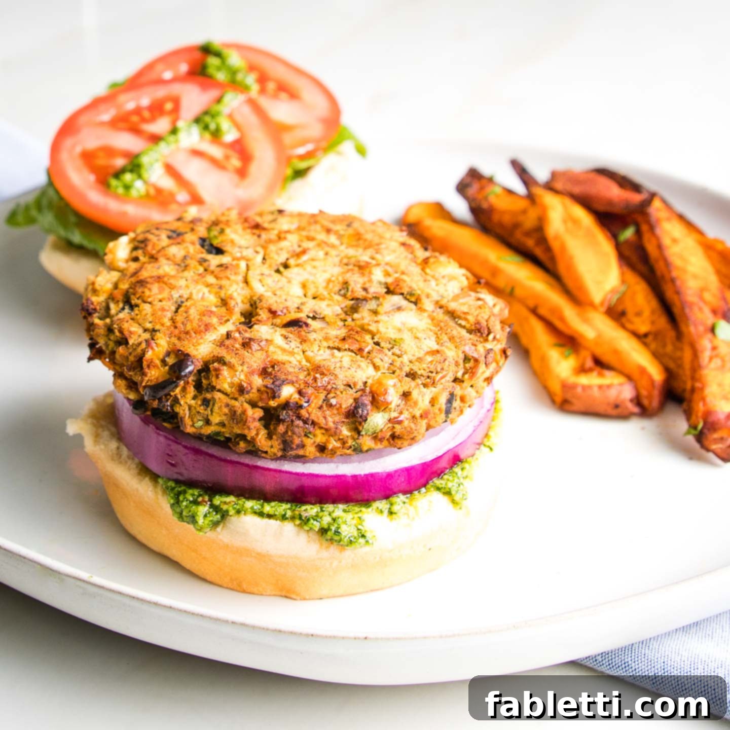 Hearty Homemade White Bean Veggie Burgers 2 An open-faced white bean veggie burger showcasing a thick patty, fresh purple onion, crisp lettuce, juicy tomato slices, and a generous dollop of green arugula aioli, served alongside a pile of crispy sweet potato fries.