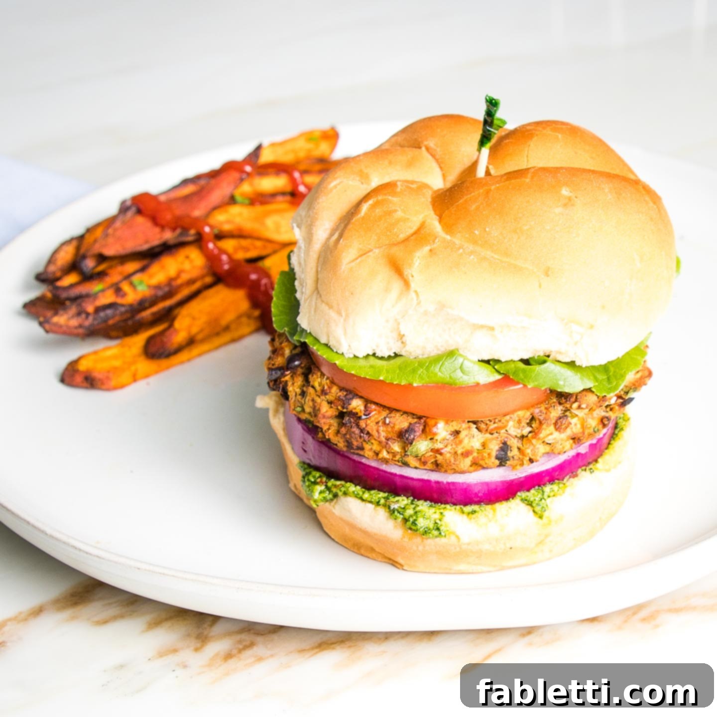 Hearty Homemade White Bean Veggie Burgers 16 A perfectly plated white bean veggie burger on a bun, adorned with vibrant arugula aioli, purple onion, crisp lettuce, and a ripe tomato. A side of golden sweet potato fries completes the wholesome meal on a round white plate.