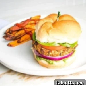 Hearty Homemade White Bean Veggie Burgers 17 Veggie burger on a bun with green arugula aioli, purple onion, lettuce and tomato on a bun with sweet potato fries on the side of a round white rimmed plate.
