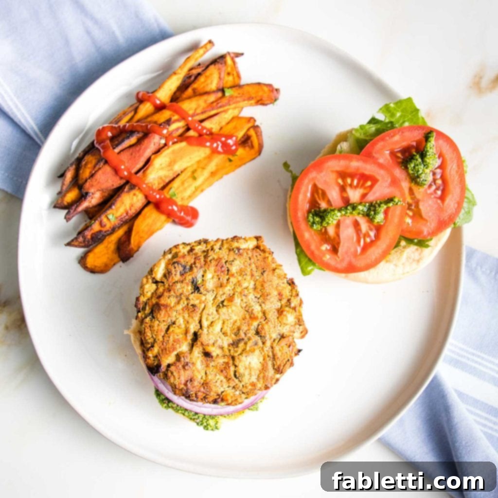 Hearty Homemade White Bean Veggie Burgers 18 A vibrant white bean burger on a toasted bun, garnished with fresh lettuce, juicy tomatoes, thinly sliced purple onion, and a generous dollop of creamy arugula aioli. A side of golden sweet potato fries, lightly drizzled with ketchup, completes this inviting meal on a round white plate.