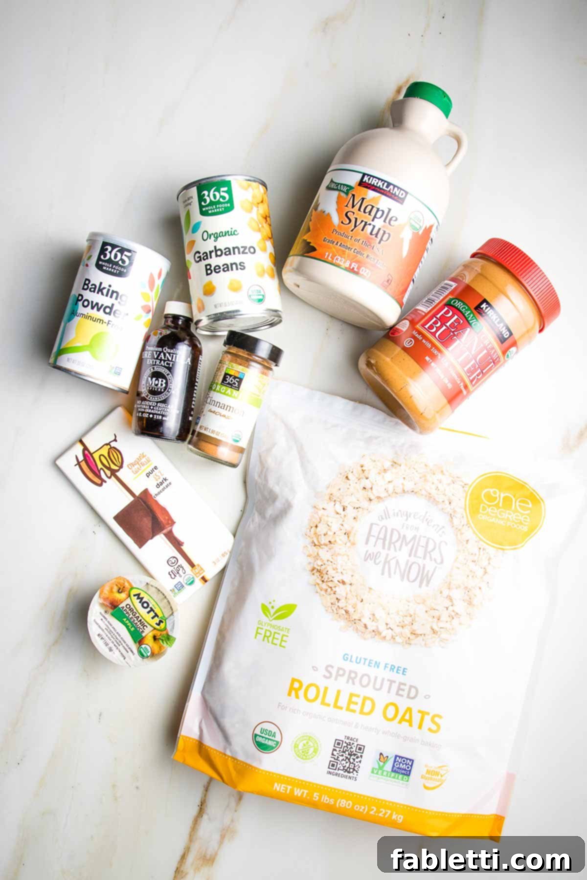 Decadent Vegan Gluten-Free Chocolate Chunk Skillet Cookie 3 A vibrant display of key ingredients including maple syrup, rolled oats, cans of chickpeas, peanut butter, cinnamon, applesauce, and a bar of dark chocolate, ready for baking.