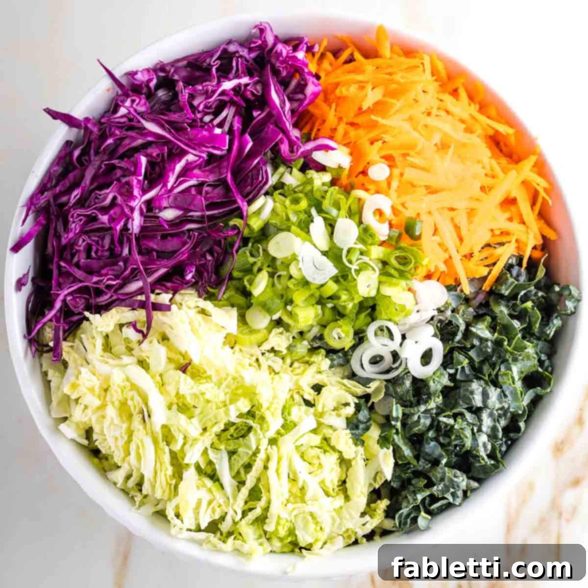 A vibrant assortment of shredded vegetables, including purple cabbage, carrots, kale, green onions, and Napa cabbage, neatly separated in sections within a large bowl.