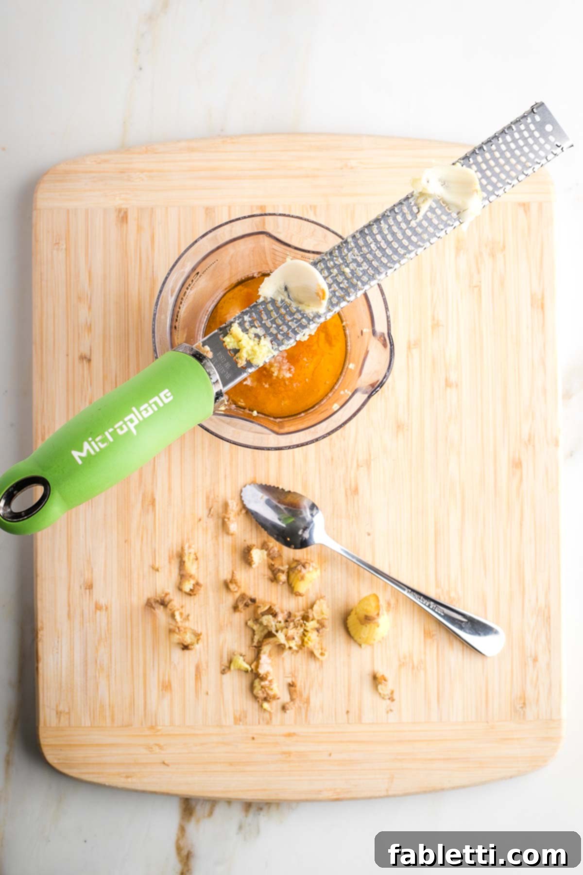 Fresh garlic being grated directly into a jar of dressing ingredients using a microplane, with ginger peels in the foreground.