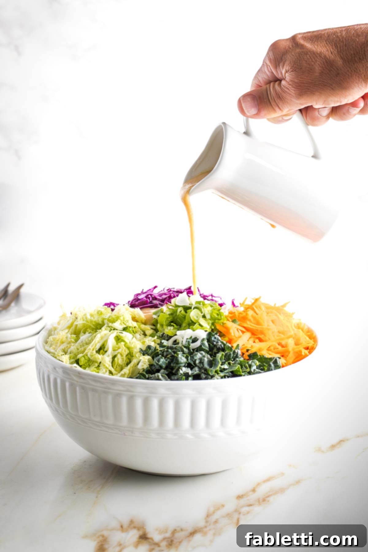 Creamy, tan-colored dressing being poured generously over a large bowl of colorful shredded vegetables.