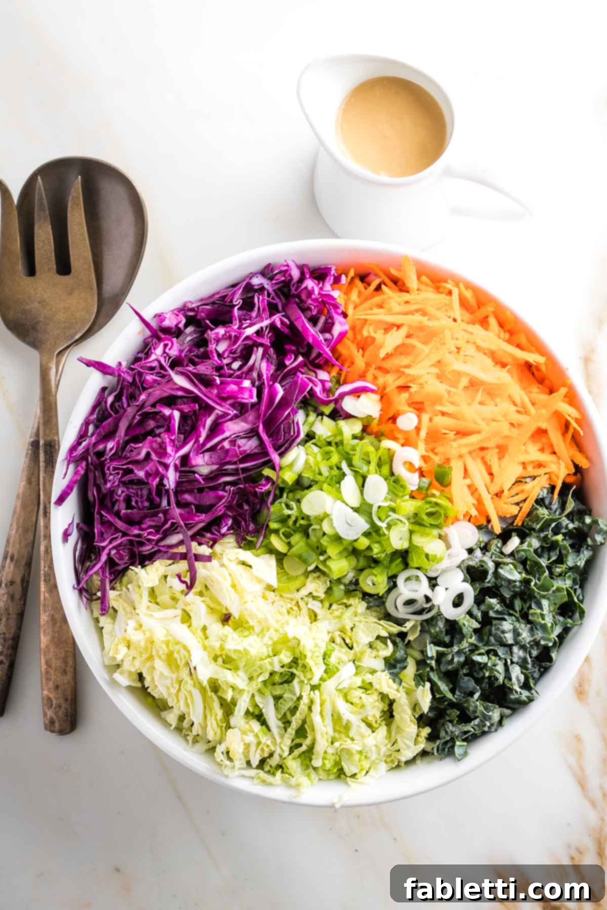 A large bowl filled with distinct sections of colorful shredded vegetables, with a small pitcher of creamy dressing placed to the side.