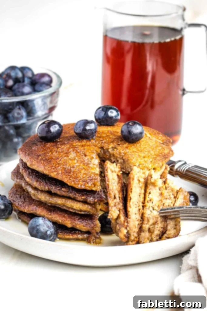Effortless Vegan Banana Oat Blender Pancakes 2 Stack of banana oat pancakes with a bite cut and ready to go on a fork. Blueberries and maple syrup in the background.
