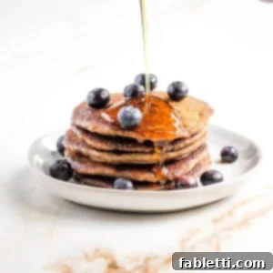 Effortless Vegan Banana Oat Blender Pancakes 9 Syrup pouring onto stack of pancakes with blueberries on top.