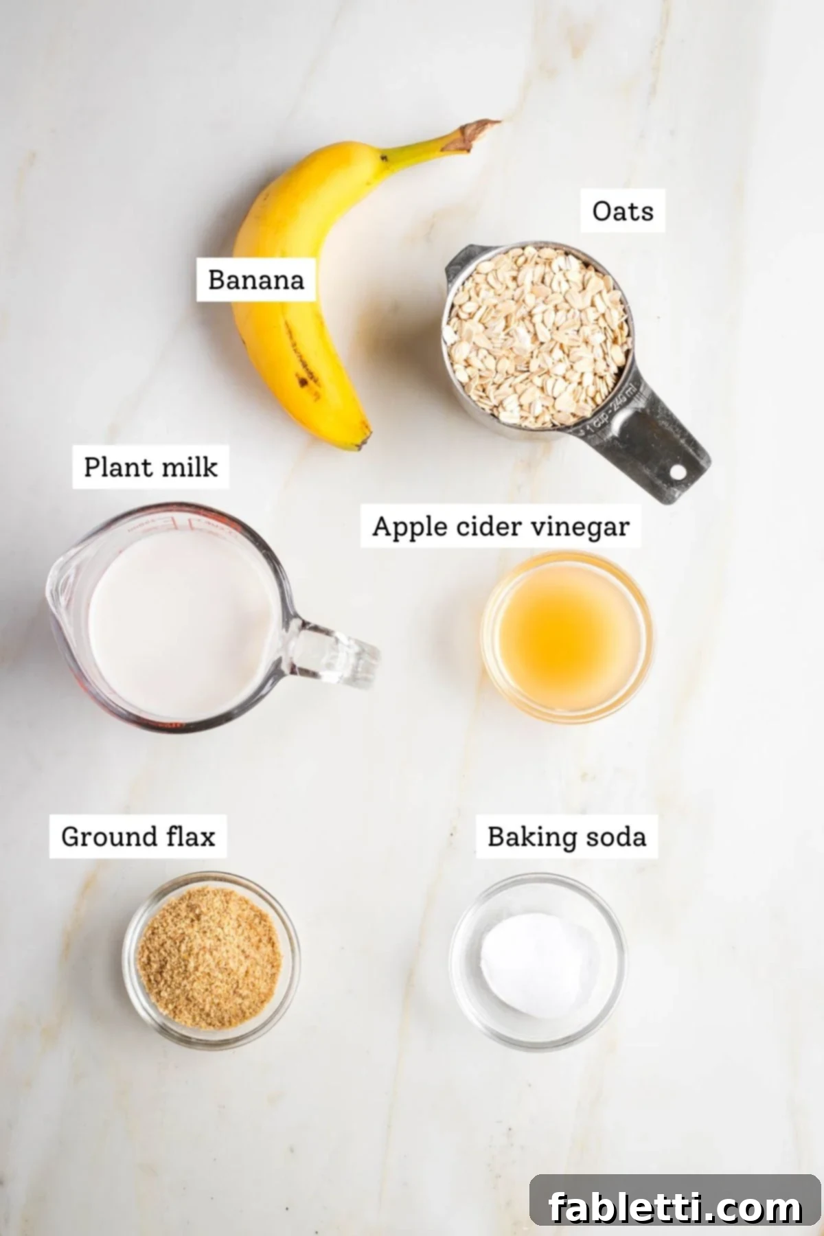 Effortless Vegan Banana Oat Blender Pancakes 3 Ingredients for vegan banana oat pancakes laid out on a table: almond milk, apple cider vinegar, ground flax, oats, bananas, and baking soda.