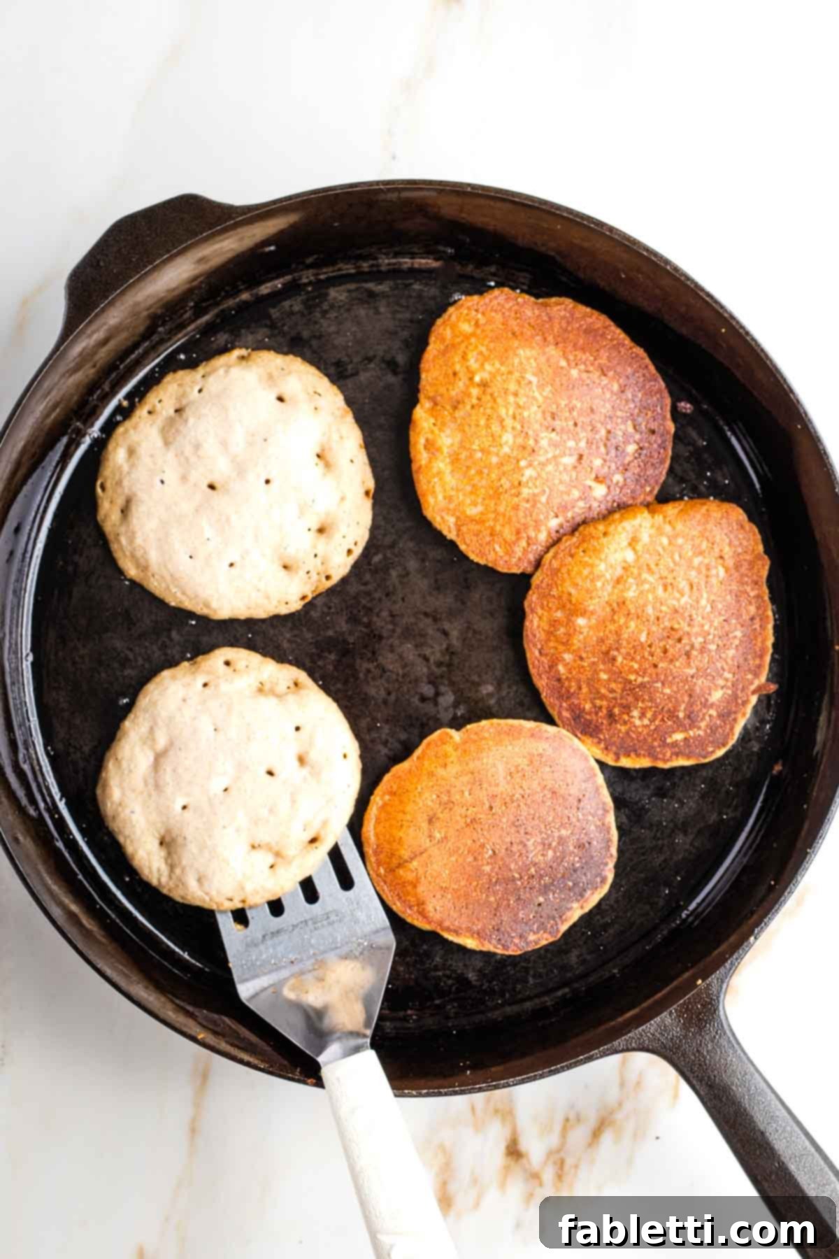 Effortless Vegan Banana Oat Blender Pancakes 6 Pancakes with bubbles, and some already flipped in a cast iron skillet.