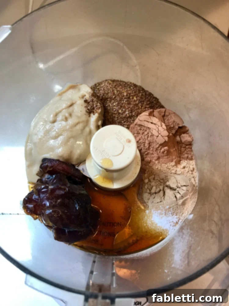 Sweet Halva Delights 4 Ingredients in the bowl of a food processor: tahini, ground flax, cacao powder, maca powder, maple syrup and dates.