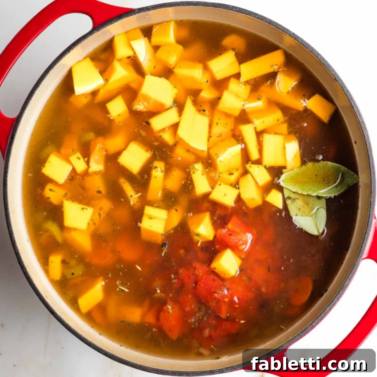 Hearty Fall Vegan Minestrone 9 Cubes of butternut squash, diced tomatoes and bay leaves, floating in broth in a dutch oven.