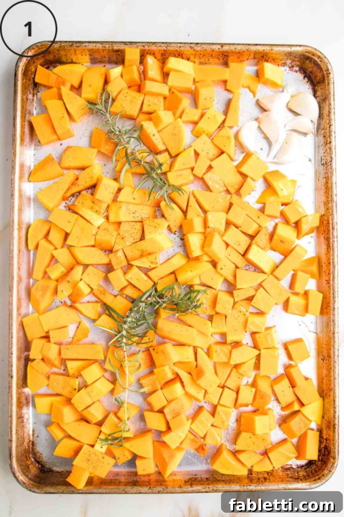 Velvety Vegan Butternut Bliss 10 Cubes of butternut squash with cloves of garlic and fresh rosemary on a large rimmed baking tray.