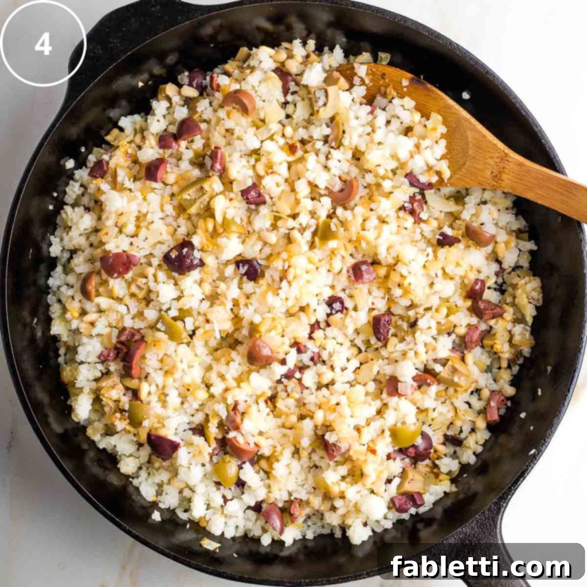 Mediterranean Cauliflower Medley 7 Step 4: Mediterranean cauliflower rice with olives and pine nuts cooking in a cast iron pan, showing steam.