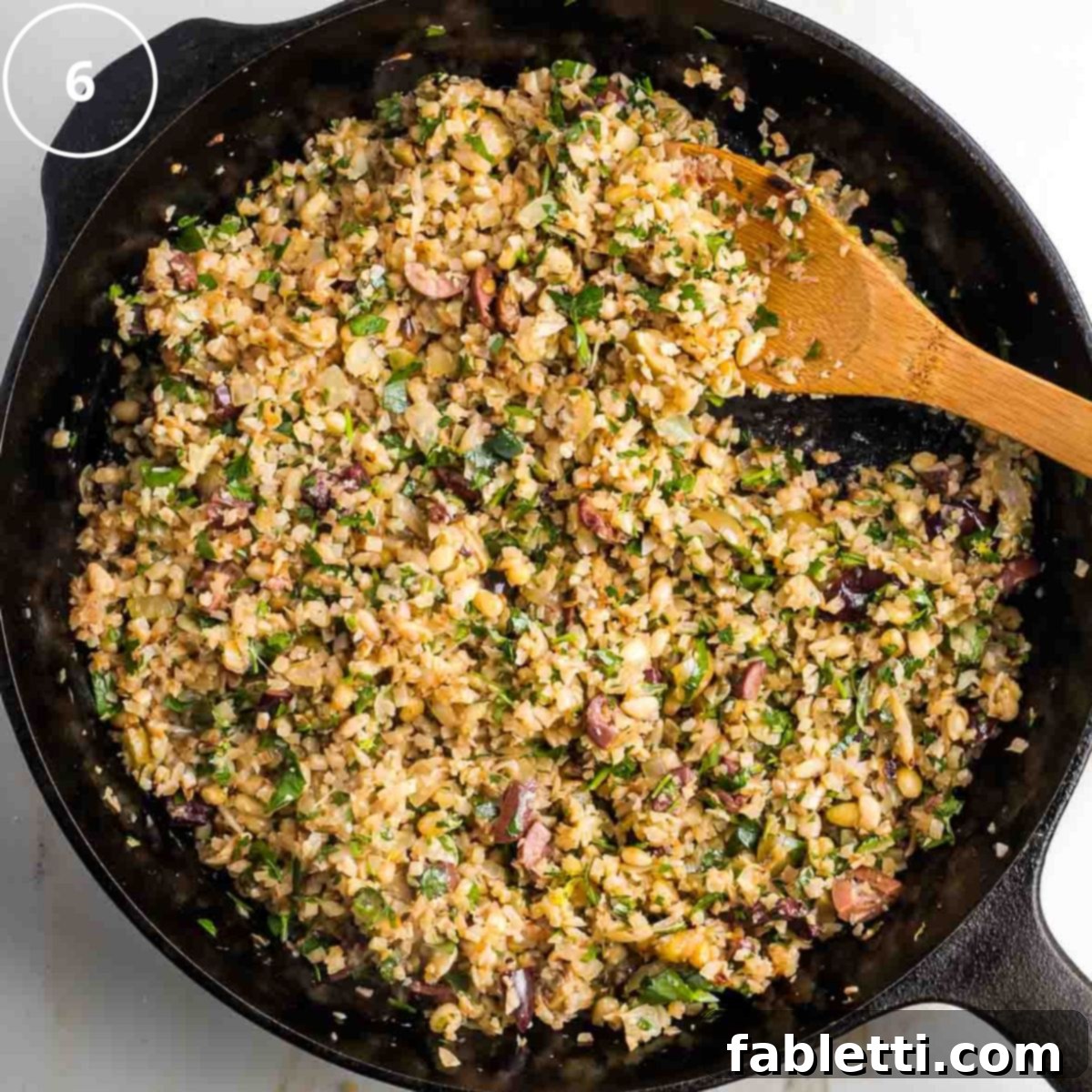 Mediterranean Cauliflower Medley 9 Step 6: Wooden spoon stirring Mediterranean cauliflower rice in a cast iron skillet with olives, fresh herbs, and pine nuts, ensuring flavors are combined.