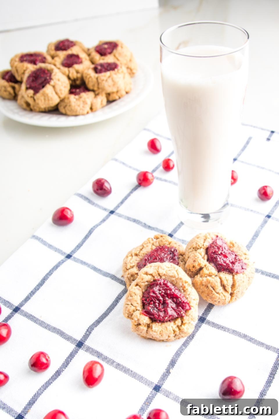 Almond Gem Cookies 3 A plate of healthy almond thumbprint cookies with cranberry chia jam, a glass of almond milk, and fresh cranberries sprinkled nearby.