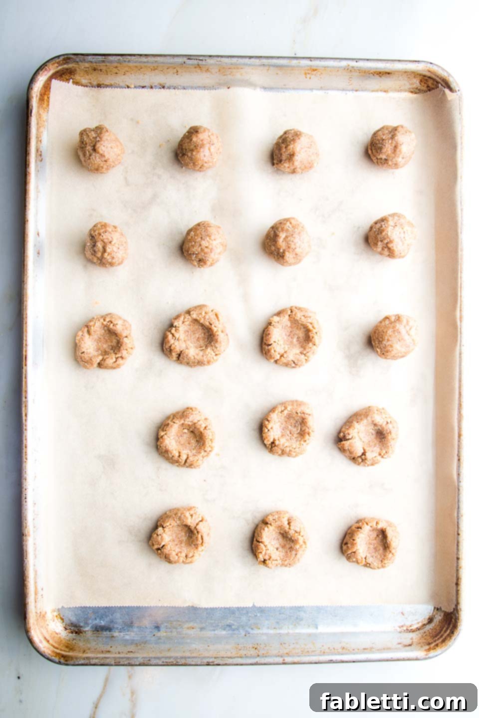 Almond Gem Cookies 7 Rows of rolled cookie dough balls on a parchment-lined baking sheet, some with thumbprint indents already made.