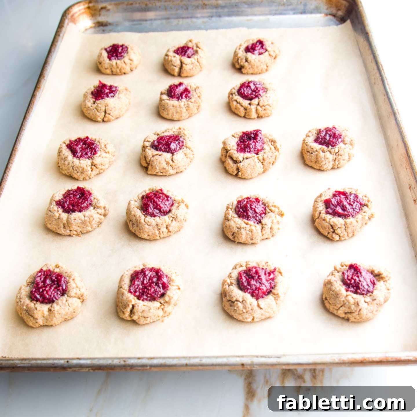 Almond Gem Cookies 9 A baking tray filled with raw thumbprint cookies, each with a vibrant cranberry chia jam filling, ready for the oven.