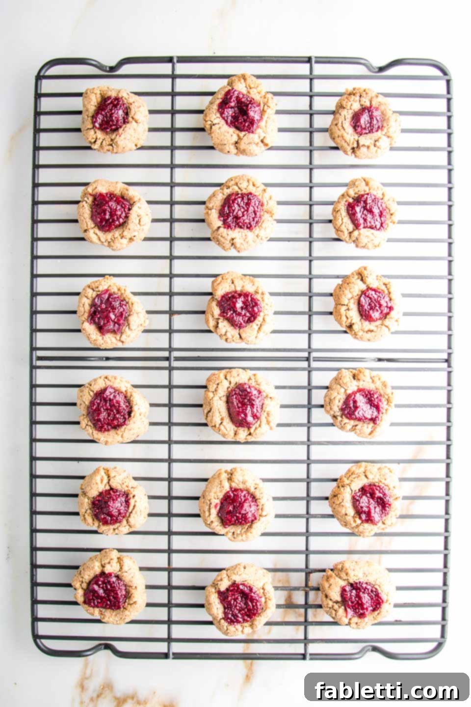 Almond Gem Cookies 10 Freshly baked thumbprint cookies, with bright red jam centers, cooling on a wire rack.