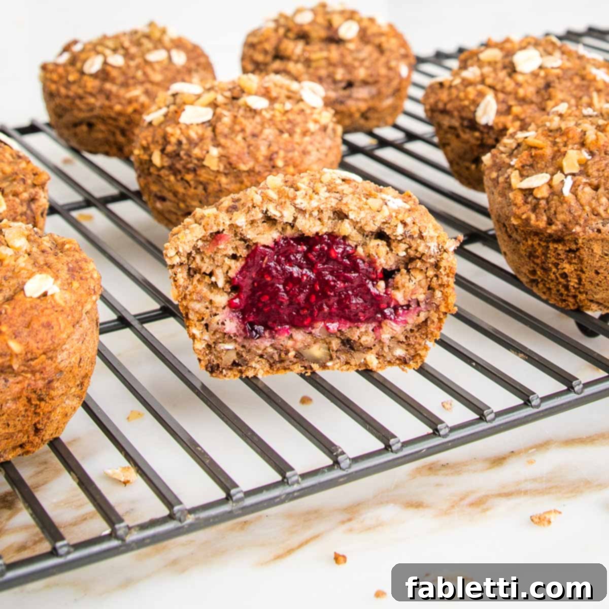 Oatmeal muffins, filled with jam, cooling on a wire rack. One muffin has been cut open and you can see the red jam inside.