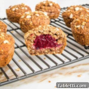Oatmeal muffins, filled with jam, cooling on a wire rack. One muffin has been cut open and you can see the red jam inside.