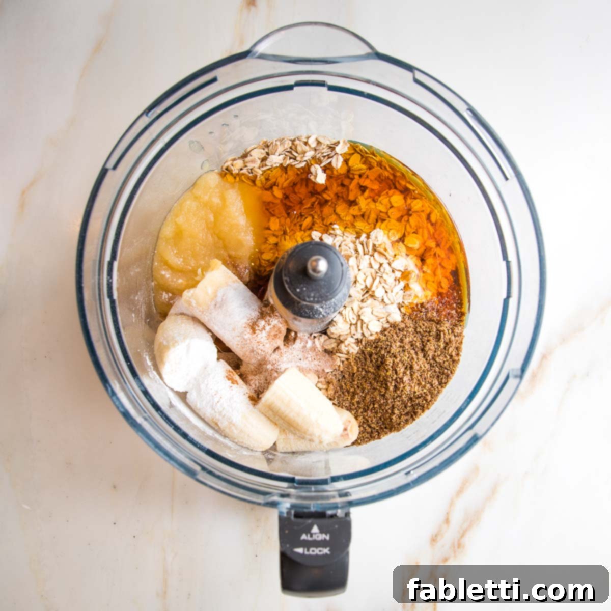 Bowl of food processor filled with bananas, applesauce, oats, flax, spices, apple cider vinegar, honey, vanilla.