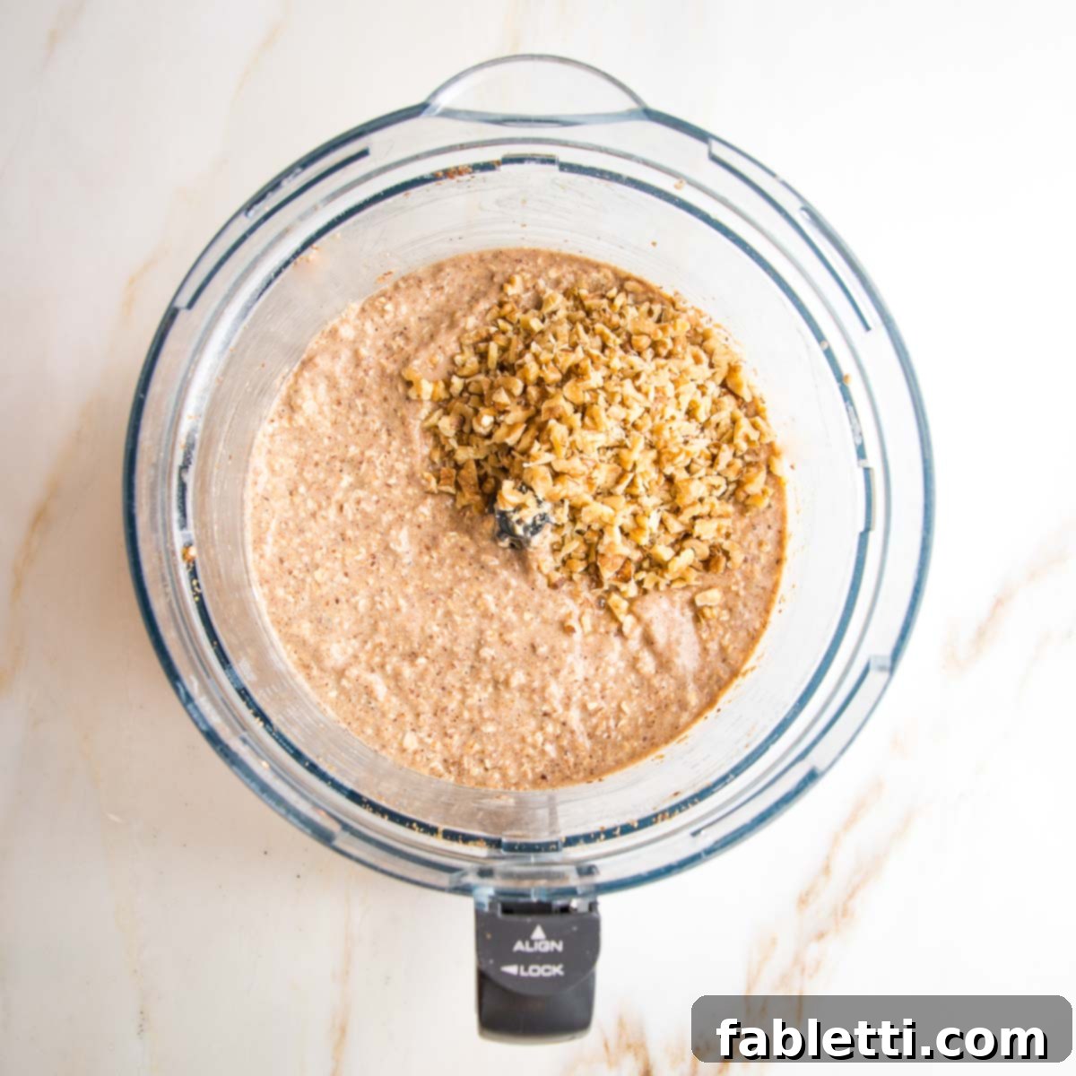Oatmeal muffin batter in the bowl of a food processor with a scoop of chopped walnuts on top.