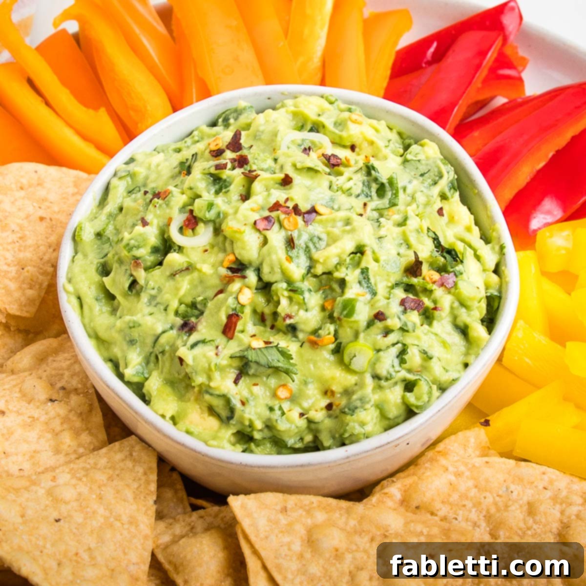 Plant-Based Super Bowl Bites 3 Close up of a bowl of chunky vegan guacamole sprinkled with red pepper flakes. The bowl is surrounded by tortilla chips and assorted color strips of bell peppers, ready for dipping.