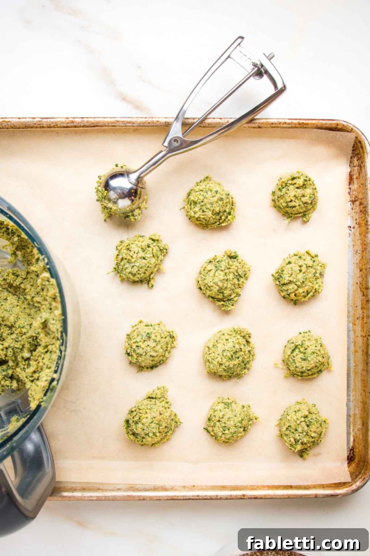 Deliciously Healthy Baked Falafel 10 A large, rimmed baking sheet lined with unbleached parchment paper, with a small scoop portioning golf-ball sized falafel mixture onto the tray, ready for baking.