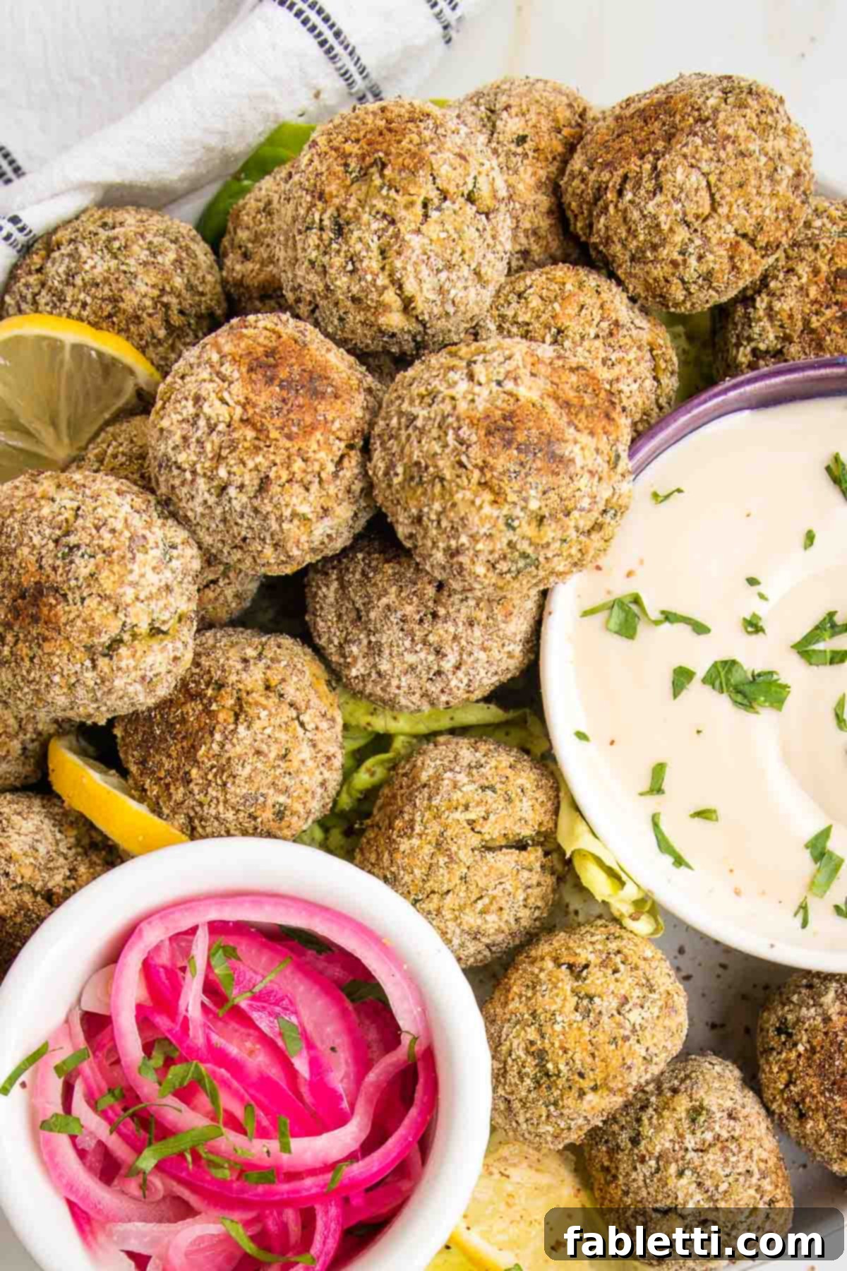 Deliciously Healthy Baked Falafel 4 A beautifully arranged plate featuring baked falafel balls, accompanied by a small white dish of vibrant red pickled onions and another dish holding a creamy tahini sauce, lightly sprinkled with fresh green herbs.