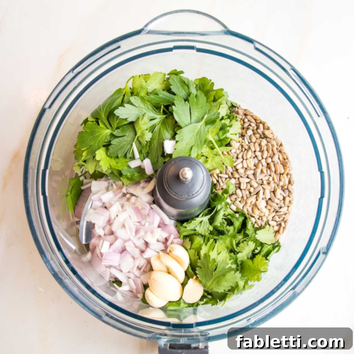 Deliciously Healthy Baked Falafel 5 A food processor bowl filled with fresh falafel ingredients including vibrant green herbs like parsley and cilantro, pungent garlic cloves, thinly sliced shallots, and raw sunflower seeds, all ready to be processed.