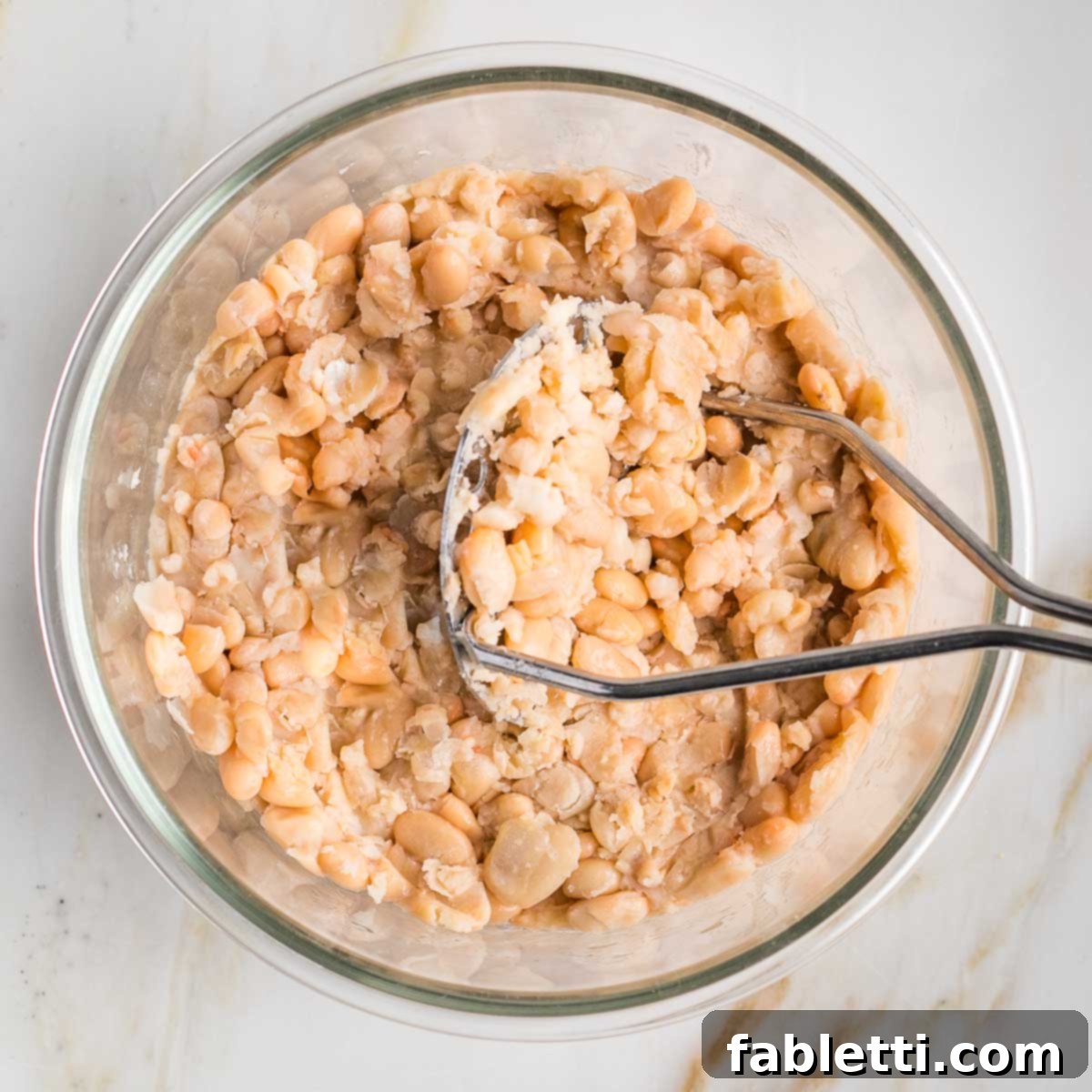 Creamy Vegan Chicken Salad 4 A potato masher mashing rinsed cannellini beans in a clear glass bowl, preparing them for vegan chicken salad.