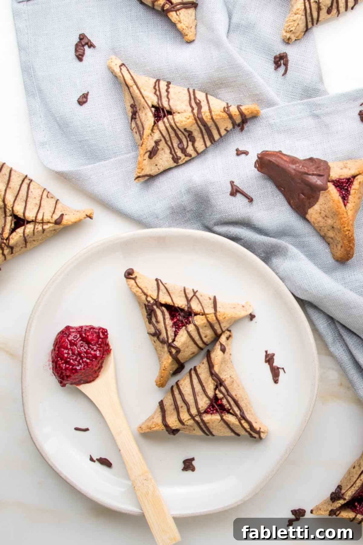 Hamentaschen cookies filled with poppyseeds or raspberry jam and drizzled with chocolate. A couple are on a white plate and the rest strewn around on a linen napkin.