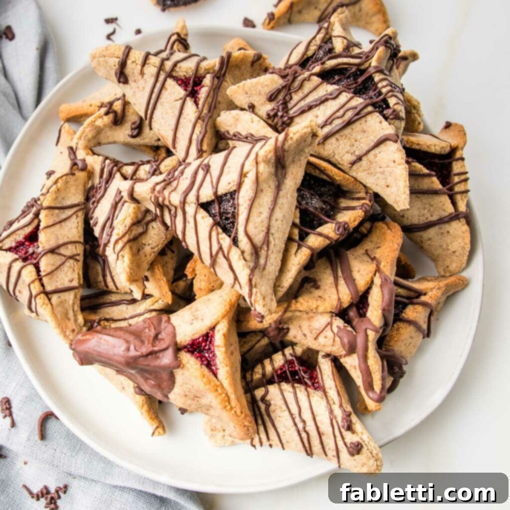 White plate filled with hamentaschen. Some filled with poppy seeds, some filled with raspberry jam. All decorated with chocolate, either dipped or swirled.