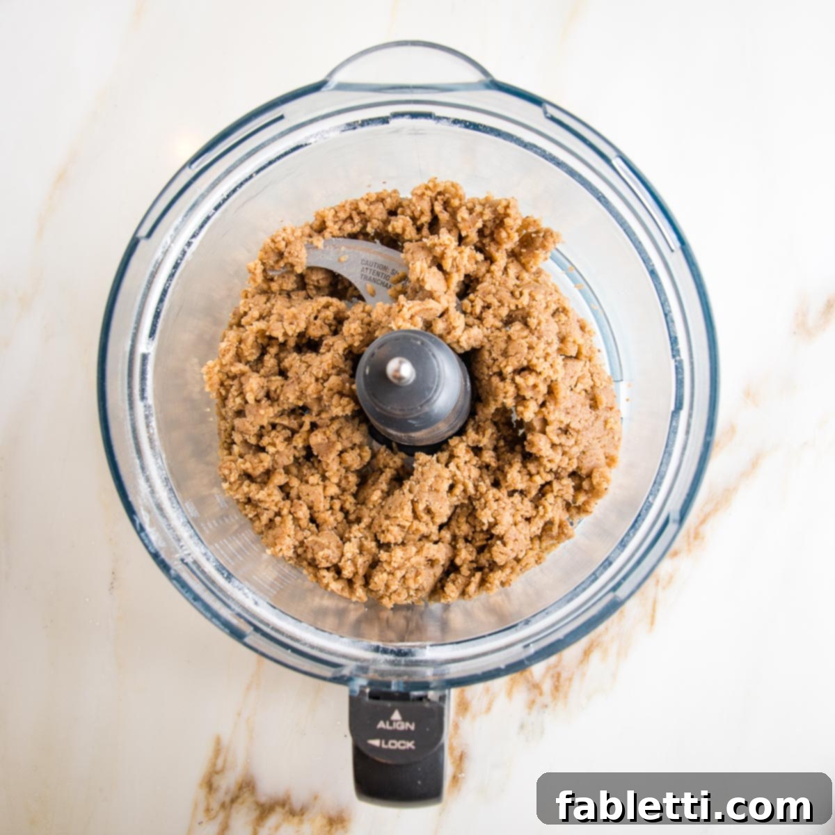 Hamantaschen dough, light brown and crumbly, in the bowl of a food processor.