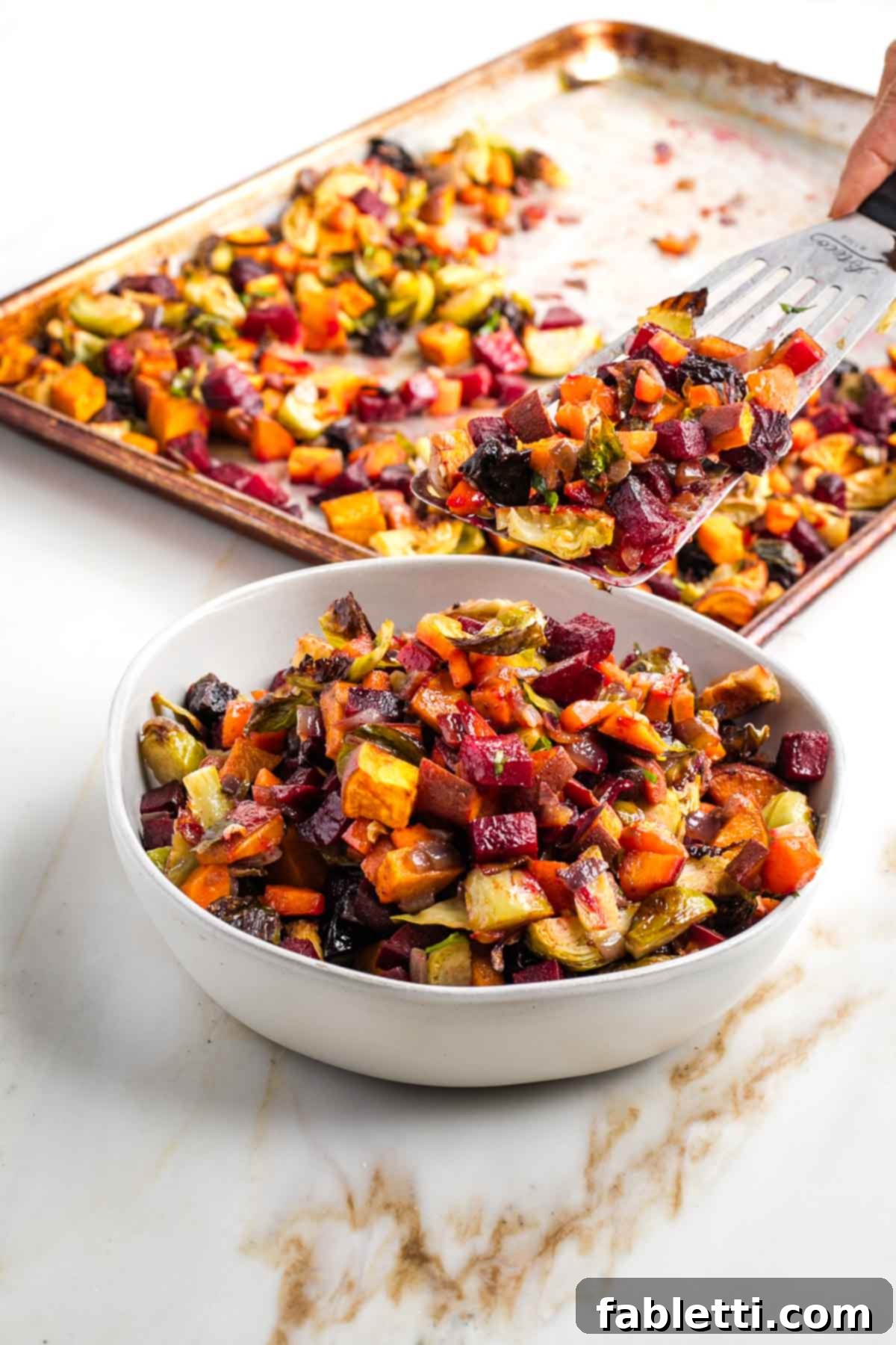 Effortless Sweet Potato and Carrot Tzimmes 8 A spatula scooping the perfectly roasted, diced root vegetables and prunes (Tzimmes) into a serving bowl, highlighting their vibrant colors and tender texture.