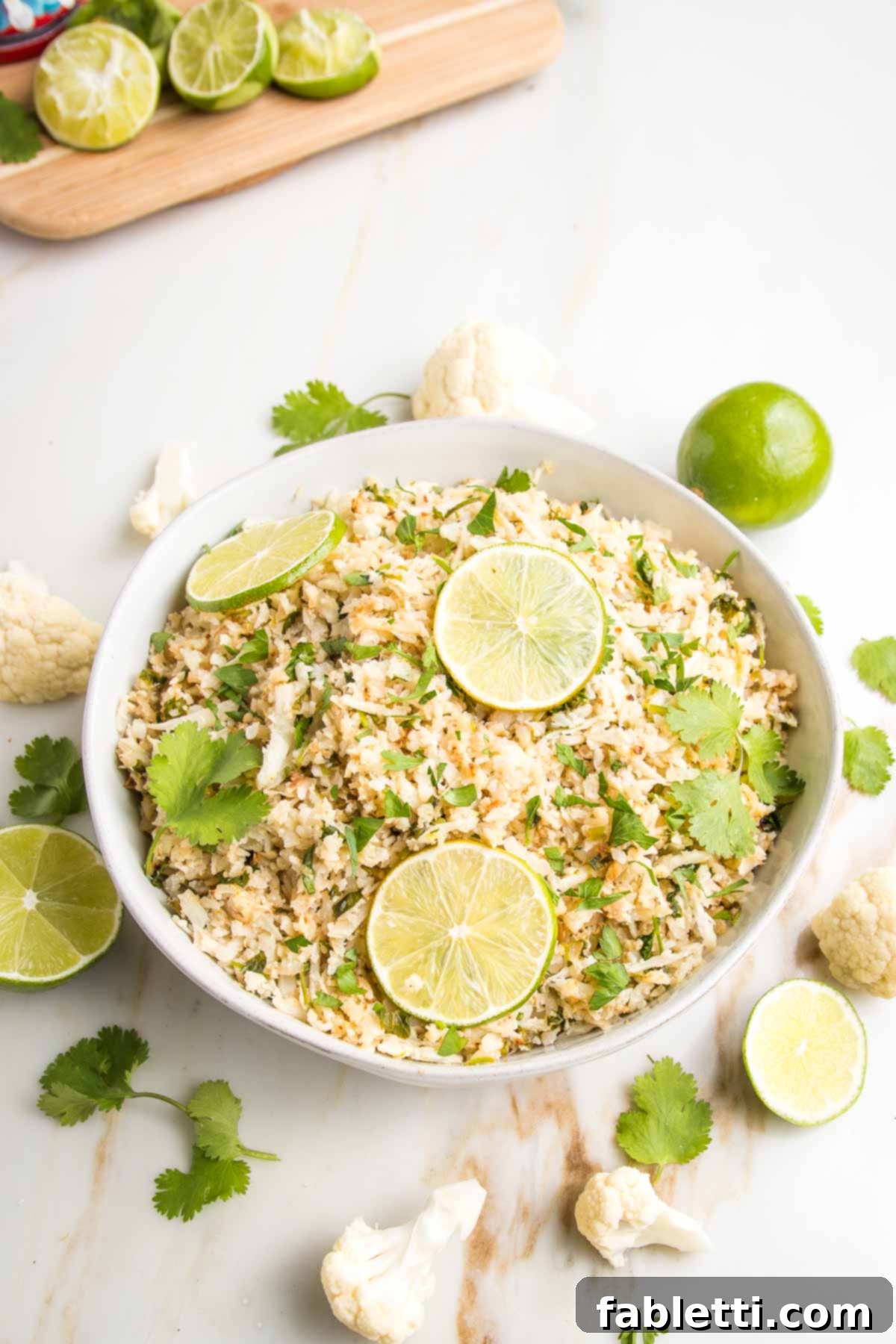 Quick Skillet Cauliflower Rice 2 Cauliflower rice in a white bowl with limes and fresh cilantro. A sprinkling of ingredients around the bowls edge.