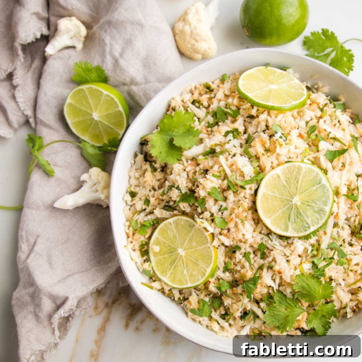 Quick Skillet Cauliflower Rice 12 White bowl filled with cauliflower rice, topped with fresh cilantro sprigs and slices of limes. A beige linnen napkin is on the side with slices of lime, florets of cauliflower and fresh cilantro.