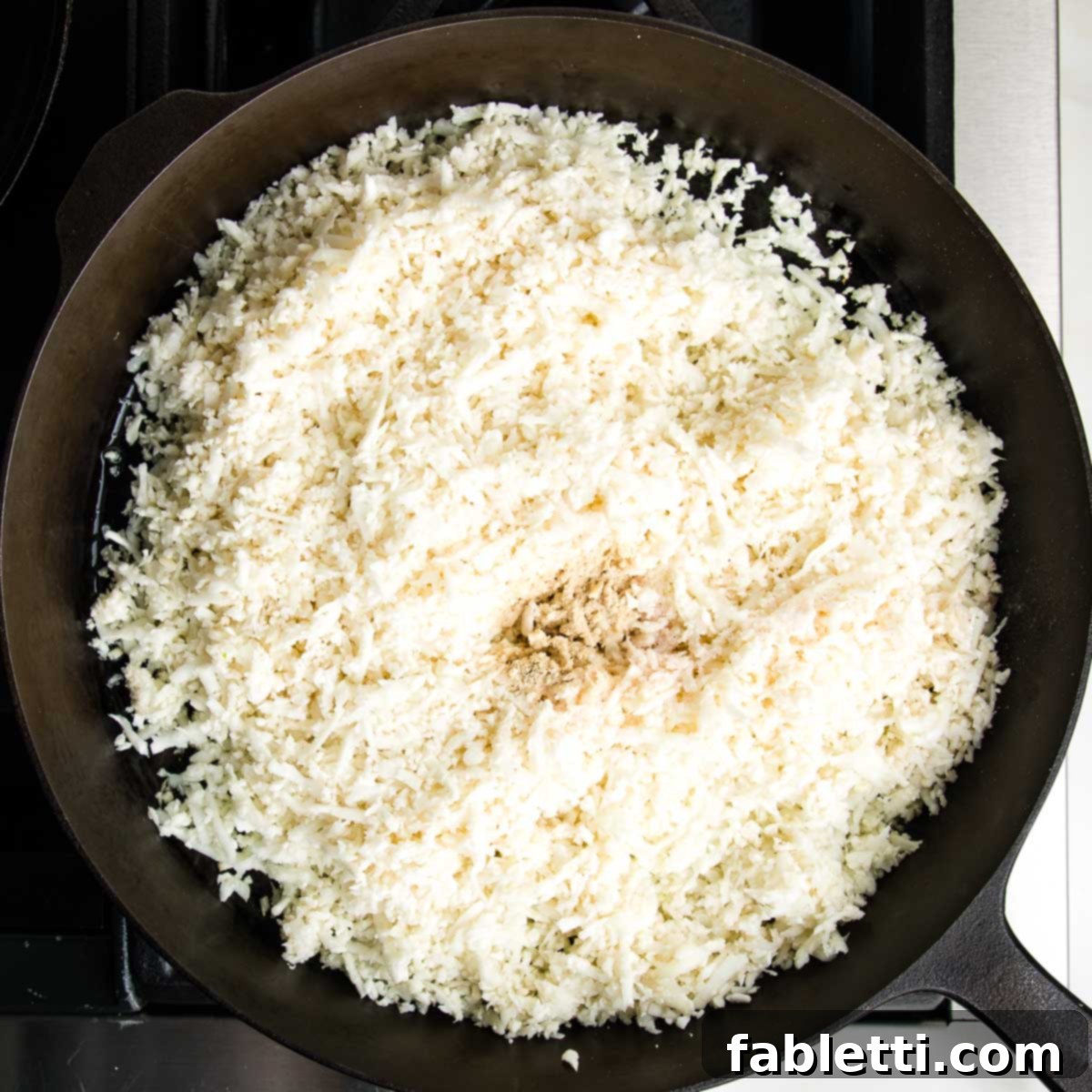 Quick Skillet Cauliflower Rice 7 Cauliflower rice in a cast iron skillet with sea salt and white pepper sprinkled into the center.