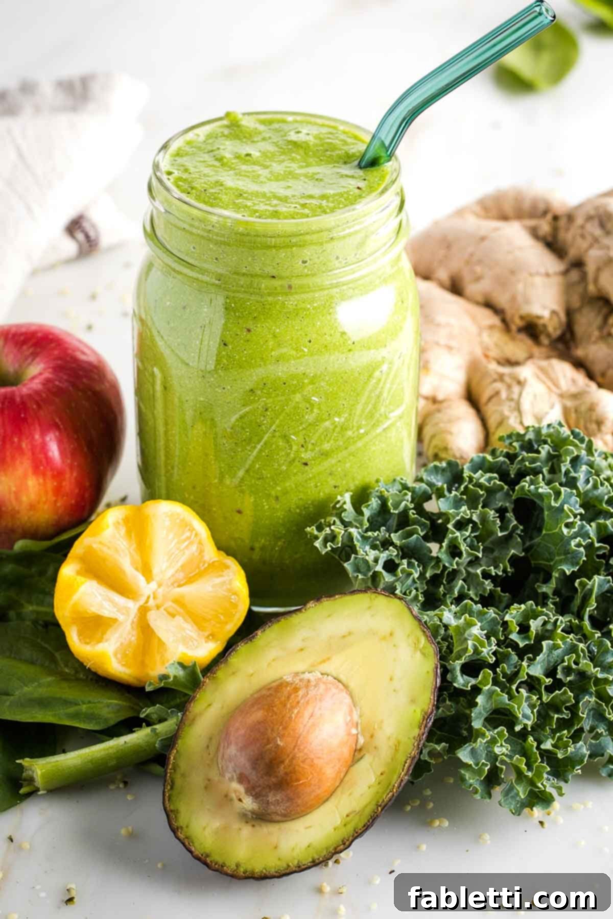 Avocado Kale Superfood Smoothie 2 A vibrant green kale avocado smoothie in a mason jar, artfully surrounded by its fresh ingredients: leafy greens, bright yellow lemon, crisp red apple, fiery ginger, and a creamy green avocado half.