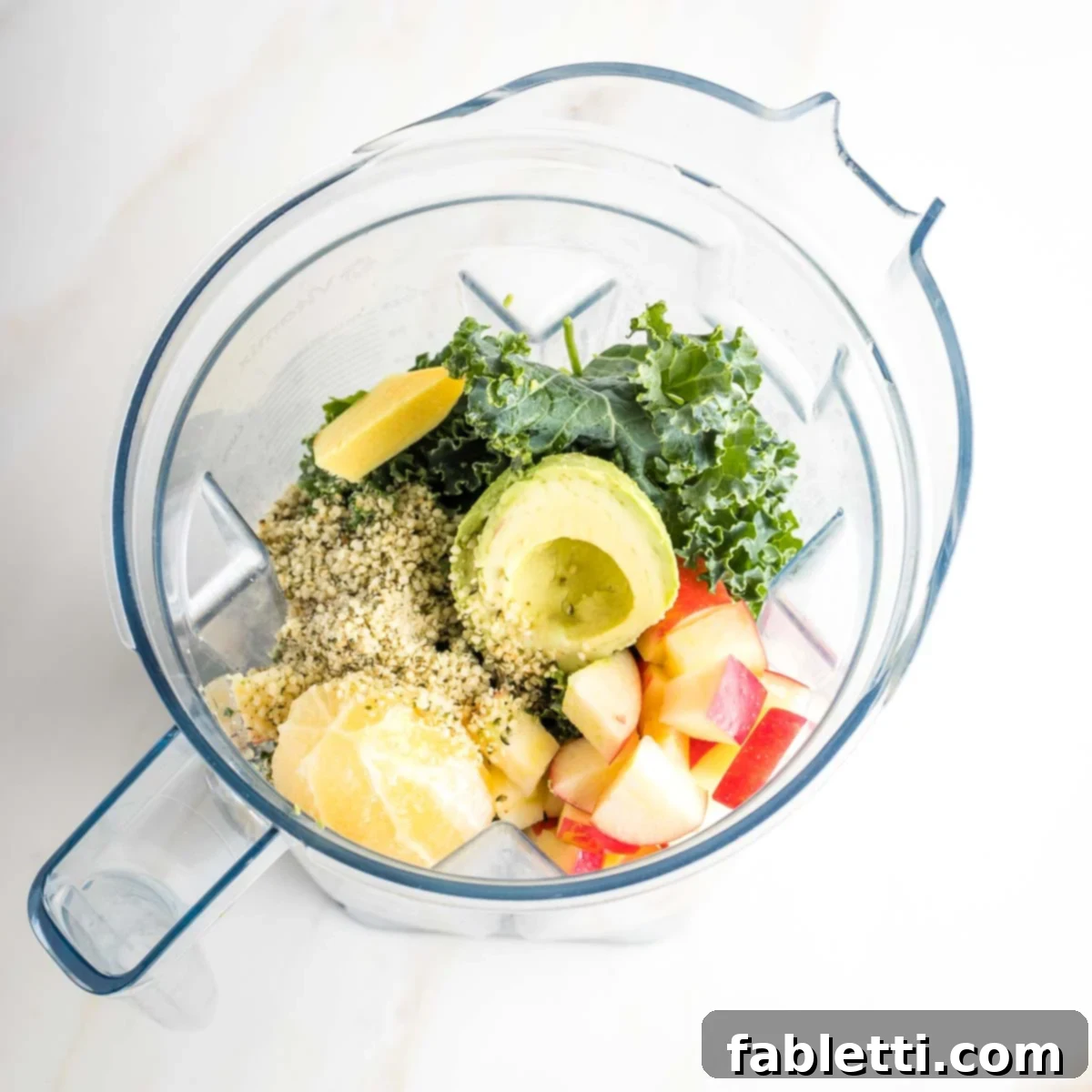 Avocado Kale Superfood Smoothie 5 A blender pitcher filled with vibrant ingredients including a halved avocado, fresh kale, spinach, lemon, ginger, apple slices, and a sprinkle of hemp seeds, ready for blending.