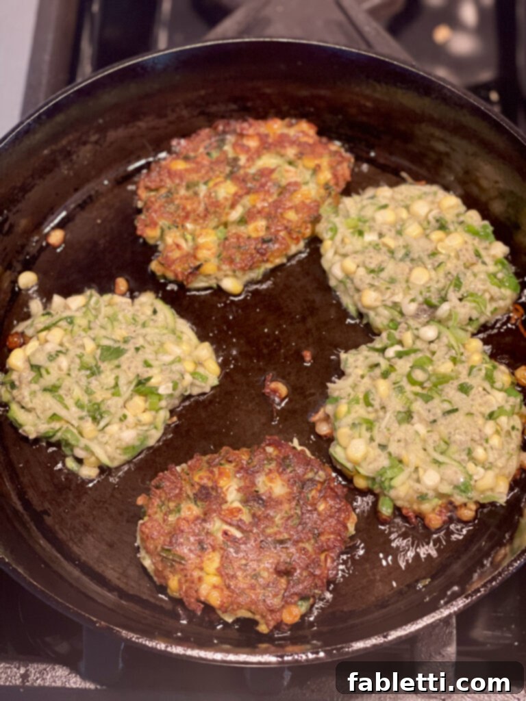 Crispy Zucchini Corn Patties 12 A large cast iron skillet on a stovetop, frying five vibrant zucchini and corn fritters. Two have already been flipped to reveal a golden-brown, crispy underside, while the remaining three show the uncooked shredded zucchini batter, awaiting their turn.