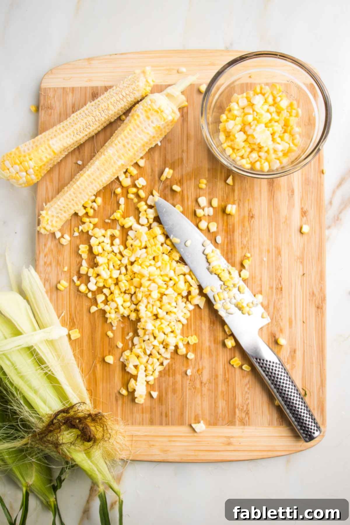Crispy Zucchini Corn Patties 6 A large cutting board showcasing corn kernels freshly cut from the cob. A small glass dish holds the bright yellow kernels, while corn husks and empty cobs are visible in the background, indicating preparation.